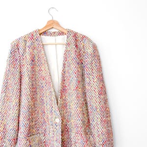 May include: A long-sleeved blazer featuring a multi-colored chevron pattern in shades of red, blue, yellow, and white. The garment has a single white button closure at the front, a collarless neckline, and a smooth white interior lining.