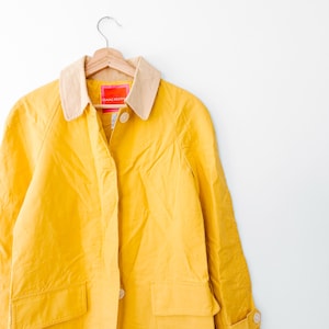 May include: A bright yellow jacket featuring a contrasting beige collar, long sleeves, and two front pockets. The garment includes a button-front closure with visible white buttons and a pink interior label with the text Isaac Mizrahi.
