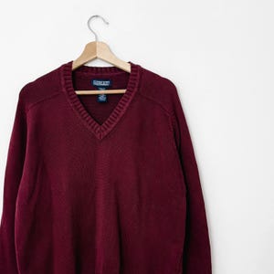 May include: A burgundy V-neck sweater hangs on a wooden hanger. The sweater has a ribbed neckline and cuffs. The Lands' End label is visible. The sweater is made of a thick knit material.