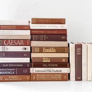 May include: A collection of hardcover books in various shades of brown, beige, and burgundy. Titles include "Caesar," "Franklin," and "The Wedding." The books are stacked in two piles, with a few standing upright.