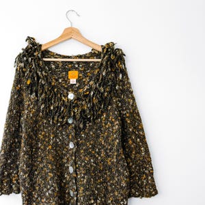 May include: A long-sleeve cardigan sweater in a chunky, multi-colored knit featuring shades of dark green, brown, and mustard yellow. The garment has a V-neckline with a thick, fringed collar, a front button closure with five round buttons, and a label.