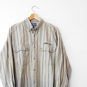 May include: A long-sleeve button-down shirt in a tan color with vertical dark blue and white stripes. The shirt features two chest pockets with button closures and the text BUGLE BOY embroidered in dark blue above the right chest pocket.
