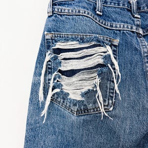 Puede incluir: Primer plano de unos vaqueros de mezclilla azules con un bolsillo desgastado. El bolsillo tiene un dise&ntilde;o deshilachado con hilos blancos, creando un aspecto &uacute;nico y desgastado. Los vaqueros cuelgan de una percha negra sobre un fondo blanco.