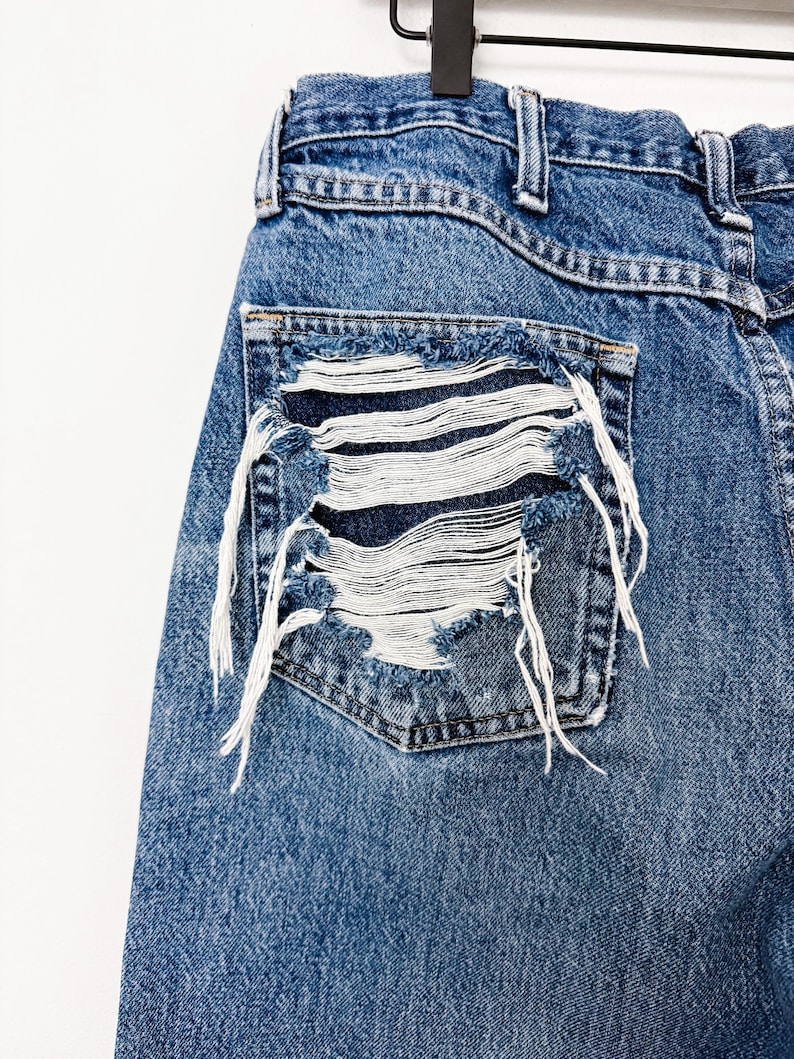 Puede incluir: Primer plano de unos vaqueros de mezclilla azules con un bolsillo desgastado. El bolsillo tiene un dise&ntilde;o deshilachado con hilos blancos, creando un aspecto &uacute;nico y desgastado. Los vaqueros cuelgan de una percha negra sobre un fondo blanco.