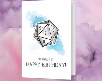 May include: A white card with a blue watercolor background and a black and white drawing of a twenty-sided die. The text on the card reads "You levelled up! Happy Birthday!"