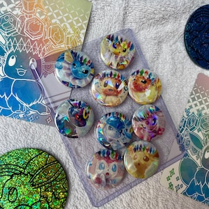 Puede incluir: Una colección de coloridos botones holográficos de personajes y tarjetas a juego. Los botones presentan varios personajes con vibrantes acentos en forma de gema. Las tarjetas tienen diseños geométricos e ilustraciones de personajes. Los artículos están dispuestos sobre una superficie blanca texturizada.