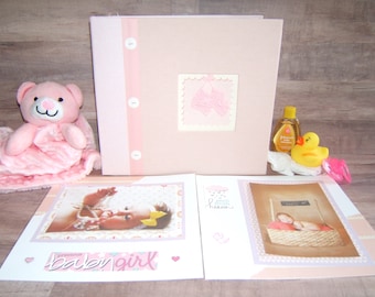 May include: Pink baby scrapbook with a pink ribbon and buttons. The scrapbook is open to reveal two photos of a baby girl. The photos are surrounded by pink and white decorative paper. The scrapbook is on a wooden surface with a pink teddy bear, a bottle of baby lotion, and a yellow rubber duck.