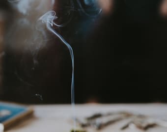 May include: A wooden incense holder with a thin incense stick burning and releasing a wisp of smoke. The holder is on a wooden surface with small pieces of dried moss scattered around it.