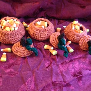 May include: Four crocheted pumpkin-shaped containers filled with candy corn. The pumpkins are orange and brown and are sitting on a pink and purple fabric surface.