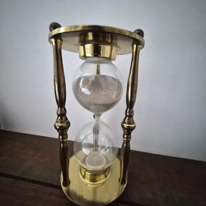 May include: An hourglass with a brass frame and clear glass bulbs. The hourglass contains sand, and the brass frame has a circular top and bottom. The hourglass is supported by four curved brass legs and two wooden finials.