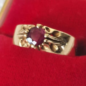 May include: A gold ring with a large, oval, deep red gemstone set in the center. The ring has decorative floral-like designs on the sides and is displayed on a red velvet surface.