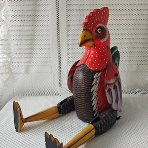May include: A hand-painted wooden rooster figurine with red, black, and white paint. The rooster is sitting with its legs outstretched and has a detailed design on its feathers.