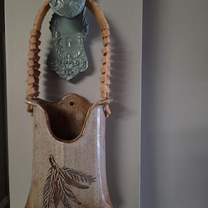 May include: A ceramic wall pocket with a speckled beige finish and a brown leaf design. The vessel features a woven light brown wicker handle and is suspended from a decorative, light blue, ornate metal wall hook with a keyhole cutout.