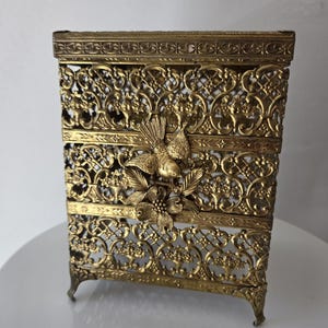 May include: A rectangular gold-toned metal box featuring an intricate openwork filigree pattern. The front panel is adorned with a three-dimensional metal bird perched above a floral motif. The box stands on four small, decorative feet.