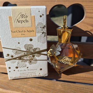 May include: A perfume bottle and box set. The box is white with gold accents and the words "Mis Arpels" and "Van Cleef & Arpels Paris." The perfume bottle is a unique shape with a gold cap and a small butterfly charm.