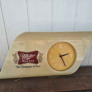 May include: Cream-colored Miller High Life clock with a round clock face. The clock features the Miller High Life logo in a maroon and gold color scheme, with the text "The Champagne of Beers".