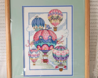 May include: Framed cross-stitch artwork featuring five colorful hot air balloons against a blue sky with white clouds. The balloons are decorated with floral patterns, hearts, and geometric designs.
