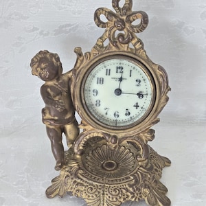 May include: Antique gold-toned clock featuring a cherub figure. The clock face is white with black numerals and hands, and the words "New Haven" are visible. The clock has an ornate, decorative base and a bow-shaped top.