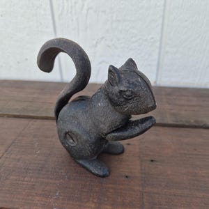 May include: A dark gray, textured cast iron squirrel figurine in a seated position with its tail curved upward. The squirrel has a visible screw on its side and is depicted holding a small object near its chest with its front paws.