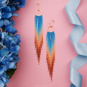 Puede incluir: Un par de pendientes largos con cuentas con un degradado de colores. Los pendientes pasan del azul al naranja y al marrón, con aros y detalles dorados. El fondo es rosa con flores azules y una cinta azul.