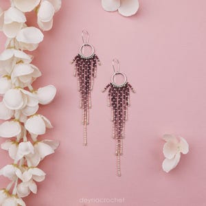 May include: A pair of long dangle earrings featuring a silver circular hoop at the top with multiple cascading strands of small beads in shades of deep plum and metallic silver. The text deynacrochet is printed at the bottom of the image.