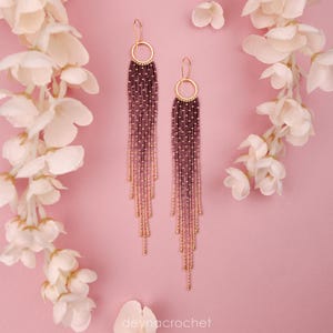 Peut inclure: Une paire de longues boucles d'oreilles à franges avec un anneau circulaire doré en haut, passant de perles violet foncé à la base de l'anneau à des perles rose clair aux extrémités, suspendues par des crochets d'oreilles dorés.