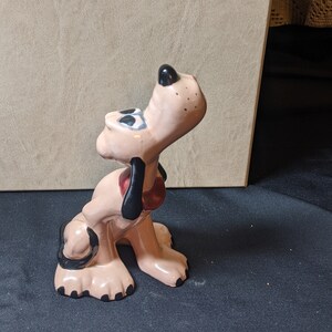 May include: A ceramic figurine of a cartoon bloodhound dog with black ears and spots. The dog is pink with a red collar and is looking up.