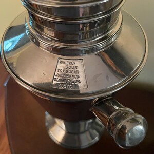 May include: A silver cocktail shaker with a rounded base and a cylindrical top. The shaker has a handle and a small rectangular plate with the words "WHISKEY SOUR TSP SUGAR JUICE LEMON GLASS RYE OR SCOTCH" engraved on it.