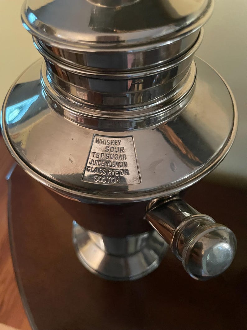 May include: A silver cocktail shaker with a rounded base and a cylindrical top. The shaker has a handle and a small rectangular plate with the words "WHISKEY SOUR TSP SUGAR JUICE LEMON GLASS RYE OR SCOTCH" engraved on it.