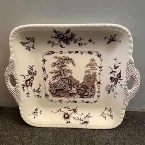 May include: A square white ceramic serving tray with brown floral and landscape designs. The tray has two handles on the sides.