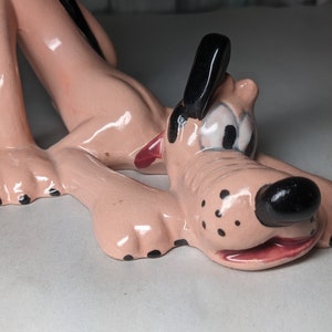 May include: A pink ceramic figurine of a cartoon dog with black ears, nose, and spots. The dog is lying on its belly with its head turned to the side.