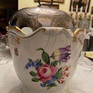 May include: White porcelain bowl with a scalloped rim and gold trim. The bowl is decorated with a floral design featuring pink roses, purple tulips, and blue wildflowers. A decorative piece for serving or display.