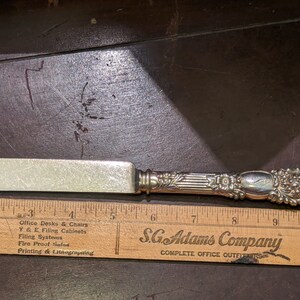 May include: A silver butter knife with an ornate handle. The handle is decorated with floral designs and has a raised inscription. The knife is 7 inches long and is resting on a wooden ruler.