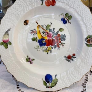 May include: White porcelain plate with a scalloped edge and a delicate lattice pattern. The plate features a central still life of colorful fruits and flowers, with additional fruit and floral accents around the rim. The rim is trimmed with gold.