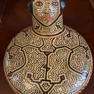 May include: A ceramic vessel with a rounded body and a cylindrical neck, featuring a face-like design. The vessel is adorned with intricate black and red geometric patterns on a cream background. The rim and neck are black.