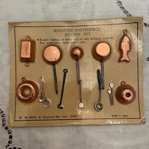 May include: A Miniature Masterpiece Kitchen Set on a tan cardboard backing. The set includes copper-colored miniature cookware, utensils, and a fish-shaped mold. Text reads: Miniature Masterpiece Kitchen Set, Exact replica in miniature, No. 6504.