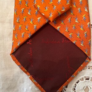 Peut inclure: Une cravate en soie orange avec un motif r&eacute;p&eacute;t&eacute; de motifs floraux stylis&eacute;s jaunes et bleus. L'int&eacute;rieur de la cravate est de couleur bordeaux fonc&eacute;, avec le nom de la marque "Salvatore Ferragamo" brod&eacute; en rouge.