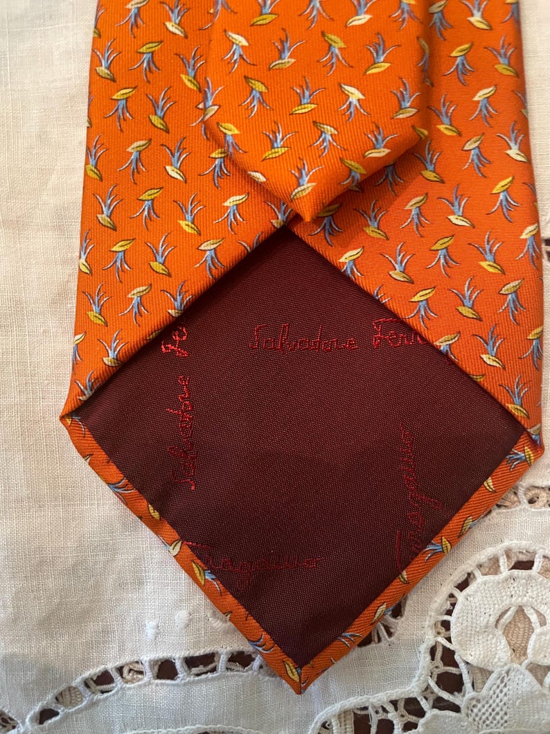 Peut inclure: Une cravate en soie orange avec un motif r&eacute;p&eacute;t&eacute; de motifs floraux stylis&eacute;s jaunes et bleus. L'int&eacute;rieur de la cravate est de couleur bordeaux fonc&eacute;, avec le nom de la marque "Salvatore Ferragamo" brod&eacute; en rouge.