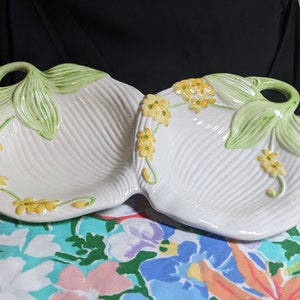 May include: White ceramic serving dish with a two-section design. The dish is decorated with yellow flowers and green leaves. The dish has a ribbed texture and a handle shaped like a leaf. A decorative piece for serving snacks or appetizers.