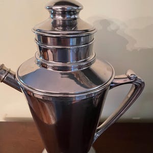 May include: A vintage silver cocktail shaker with a conical body, a lid, and a handle. The shaker has a shiny, reflective surface and a classic design, suitable for mixing drinks.