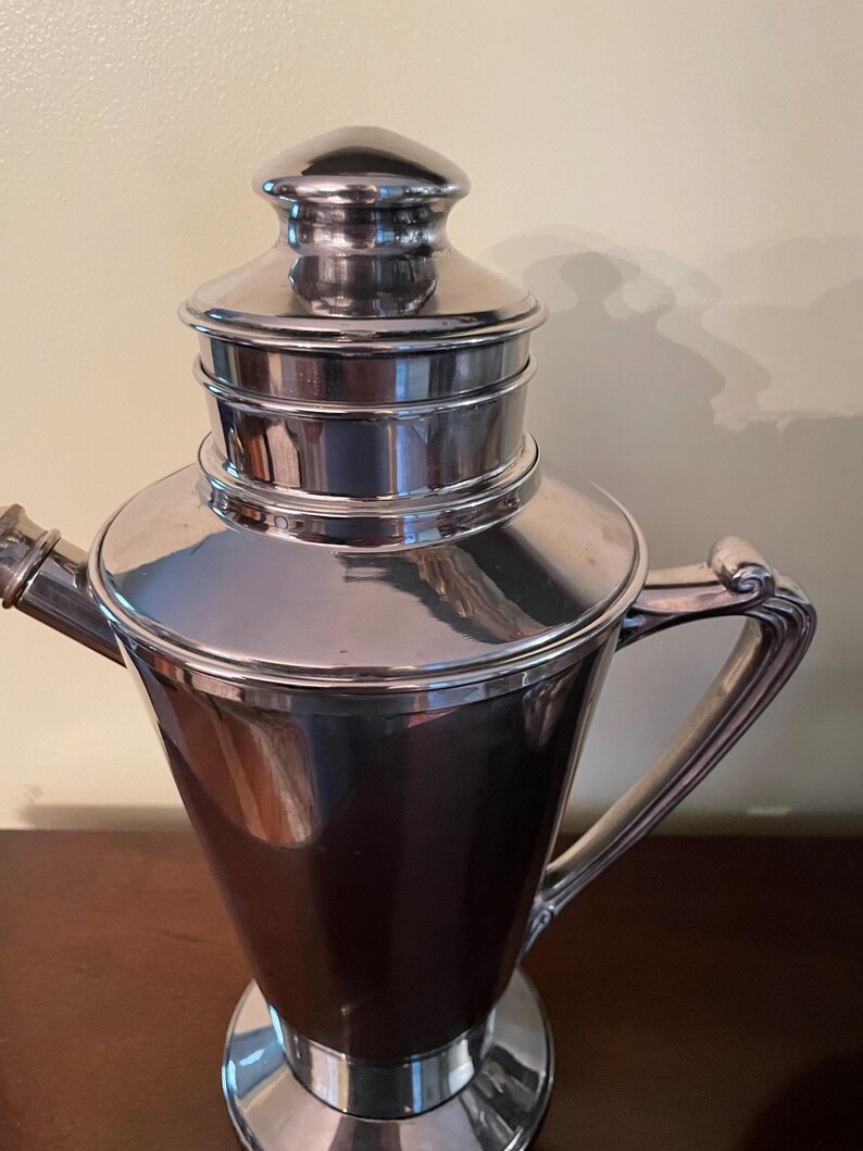 May include: A vintage silver cocktail shaker with a conical body, a lid, and a handle. The shaker has a shiny, reflective surface and a classic design, suitable for mixing drinks.