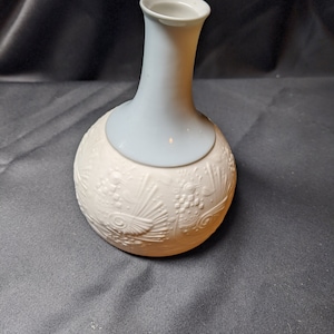 May include: A white ceramic vase with a blue glazed neck and a raised embossed design. The vase has a round body and a narrow neck.