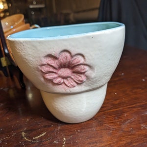 May include: A white ceramic planter with a light blue glaze and a pink flower design. The planter has a textured surface and a rounded shape.