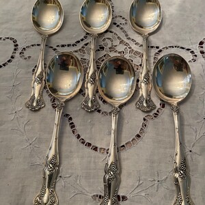 May include: Six silver spoons with ornate handles and oval bowls are arranged on a light-colored surface. The handles feature a detailed grape and leaf design. The spoons are reflective, showing the surrounding environment.