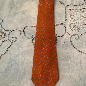 Peut inclure: Une cravate orange avec un motif r&eacute;p&eacute;t&eacute; de petites formes stylis&eacute;es en forme de feuilles dans des tons de bleu, gris et blanc. La cravate est faite d'un tissu lisse et est pr&eacute;sent&eacute;e sur une surface blanche textur&eacute;e.