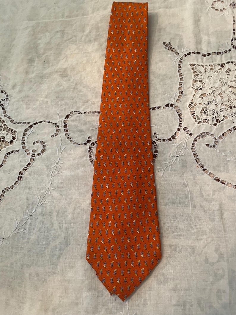 Peut inclure: Une cravate orange avec un motif r&eacute;p&eacute;t&eacute; de petites formes stylis&eacute;es en forme de feuilles dans des tons de bleu, gris et blanc. La cravate est faite d'un tissu lisse et est pr&eacute;sent&eacute;e sur une surface blanche textur&eacute;e.