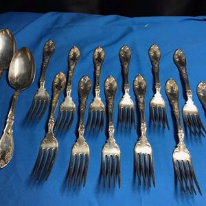 May include: A set of 11 silver-plated forks and spoons with ornate designs. The forks have three tines and the spoons have a rounded bowl. The silverware is arranged on a blue surface.