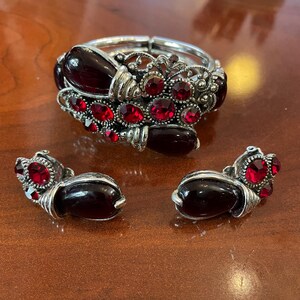 May include: A silver-toned bracelet and matching earrings set. The bracelet features a series of red, oval-shaped gemstones and smaller red stones. The earrings have a similar design, with a large red stone and smaller red stones.