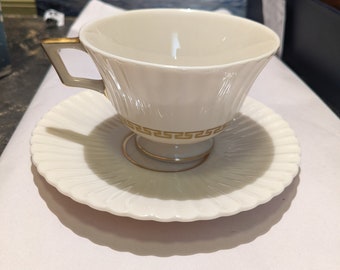May include: A white teacup and saucer with a gold trim. The teacup has a fluted design and the saucer has a scalloped edge.