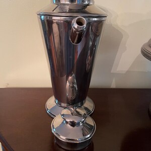 May include: A polished silver cocktail shaker with a conical body, a spout, and a lid. The shaker is displayed on a dark wooden surface. The lid and base are circular, and a small cap sits nearby. The design is vintage.